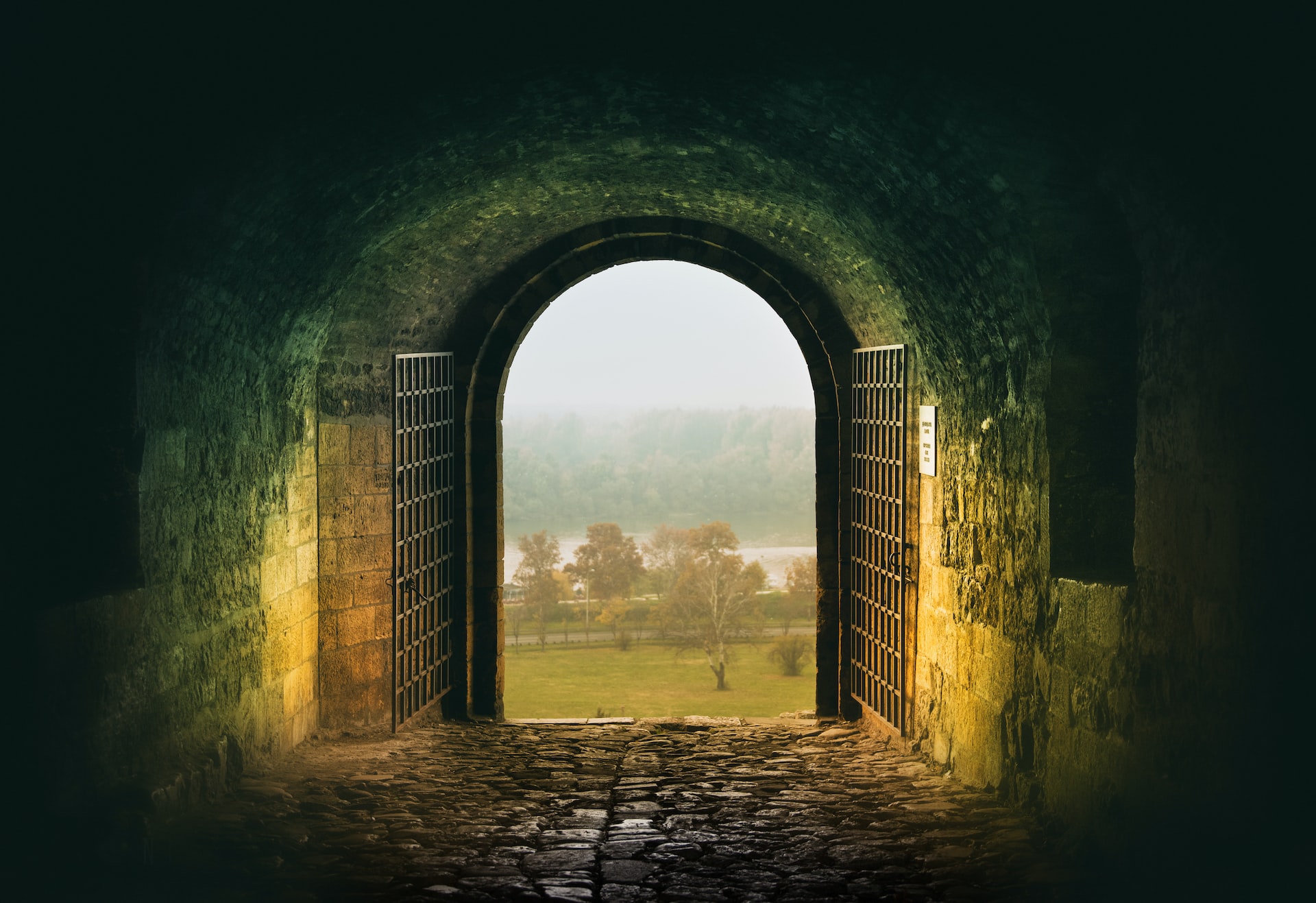 Photograph of a foggy pasture framed by a large archway with iron gates. The interior is dimly lit with hues of gold and teal, revealing a well worn cobblestone floor leading to the archway. This photograph was taken at Kalemegdan Fortress Beograd, Serbia by Nikola Knezevic.